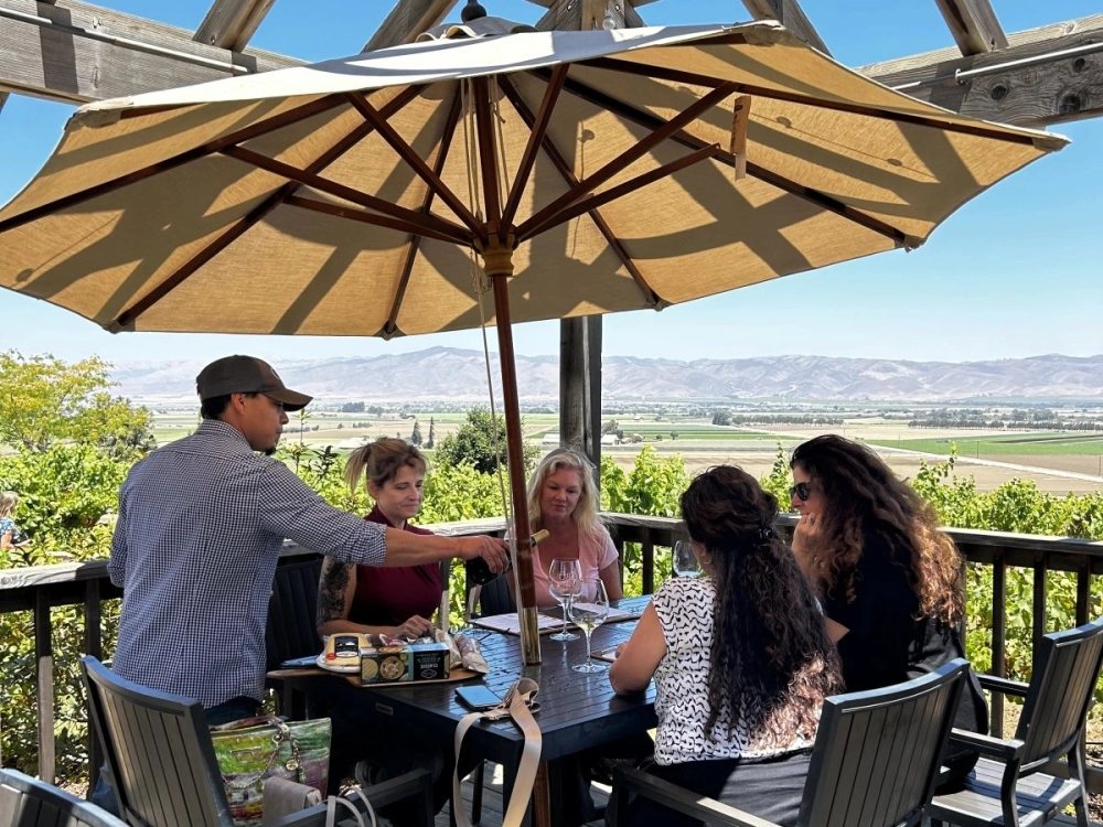 Wine tasting guests seated around a table under an umbrella on a deck, with a host pouring wine and views over the valley