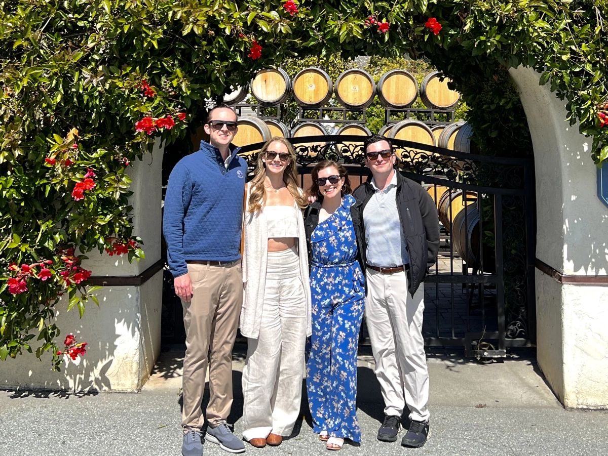 Four smiling wine tour guests standing together in front of a flowering archway and wine barrels at a Monterey County winery