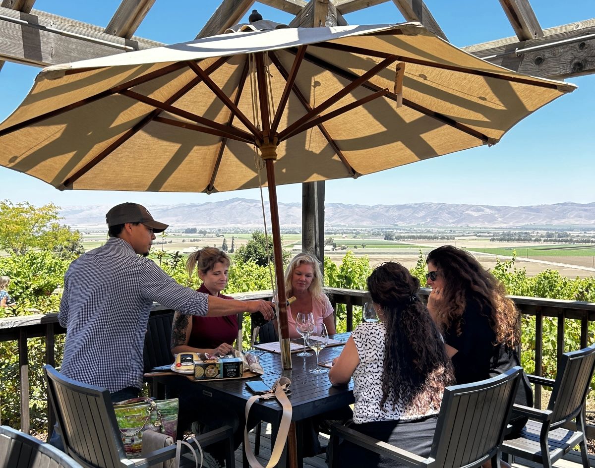 Wine tasting guests seated around a table on a deck with a host pouring wine and views over the Salinas Valley in Monterey County