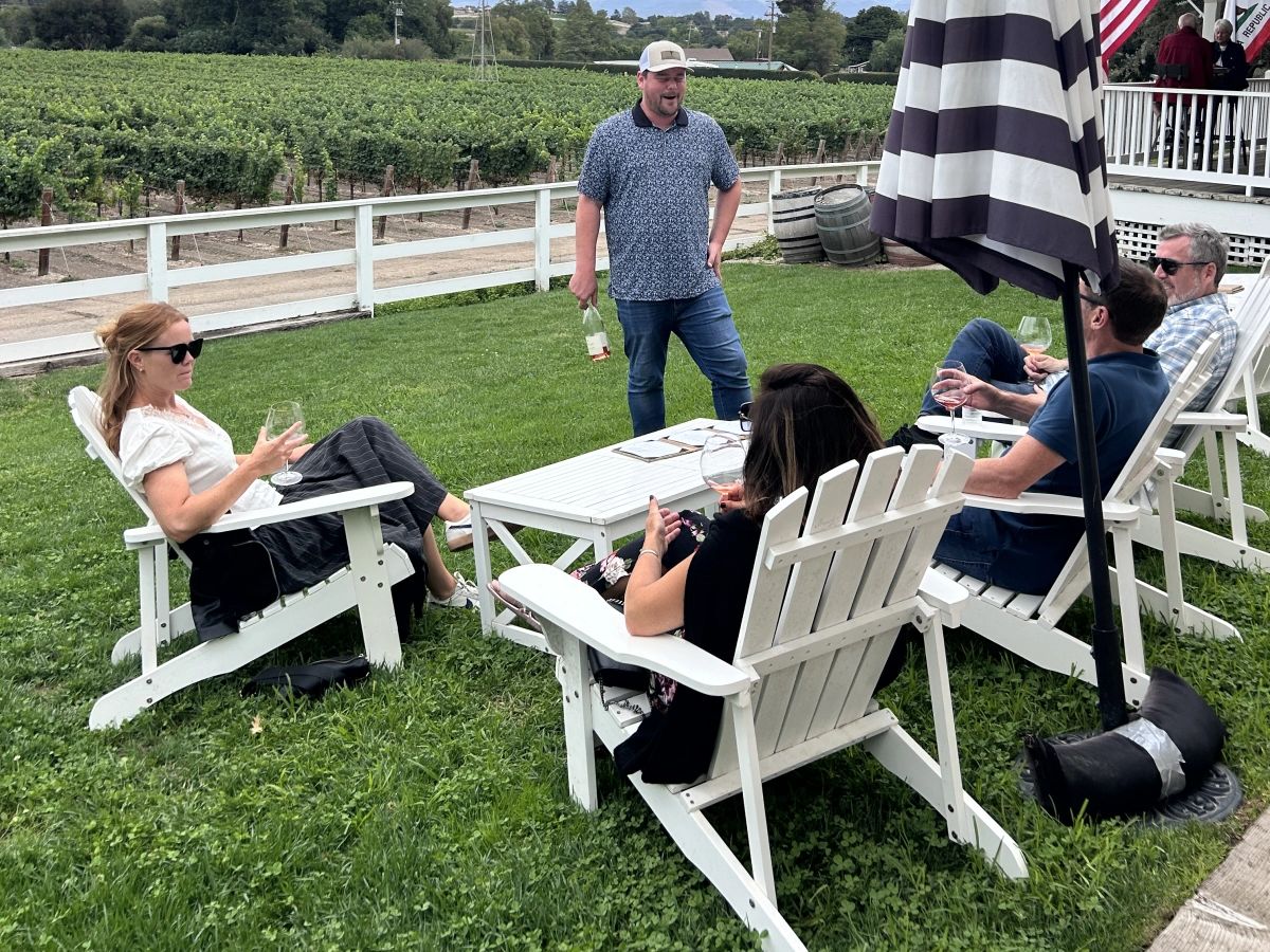 Wine tasting guests relaxing in white Adirondack chairs on a lawn beside vineyard rows while a host pours rosé in the Santa Ynez Valley